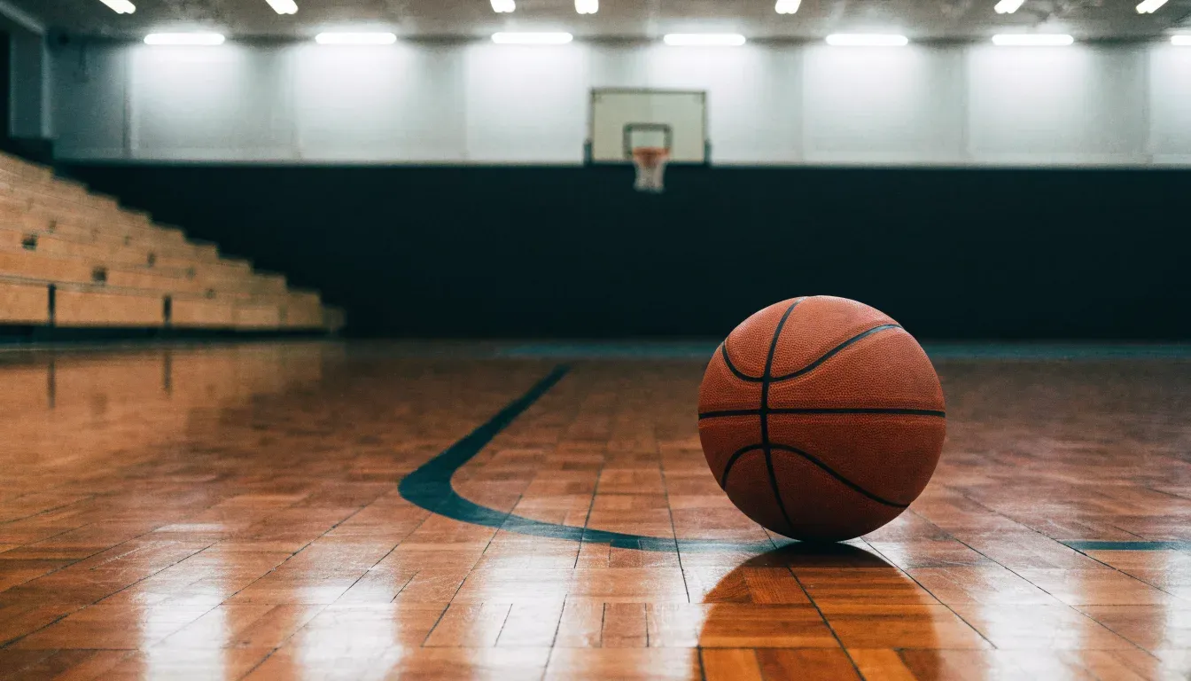 Ein Basketball liegt auf dem Parkett einer leeren Halle, von den Deckenlampen fällt gleichmässiges Licht auf den Boden.