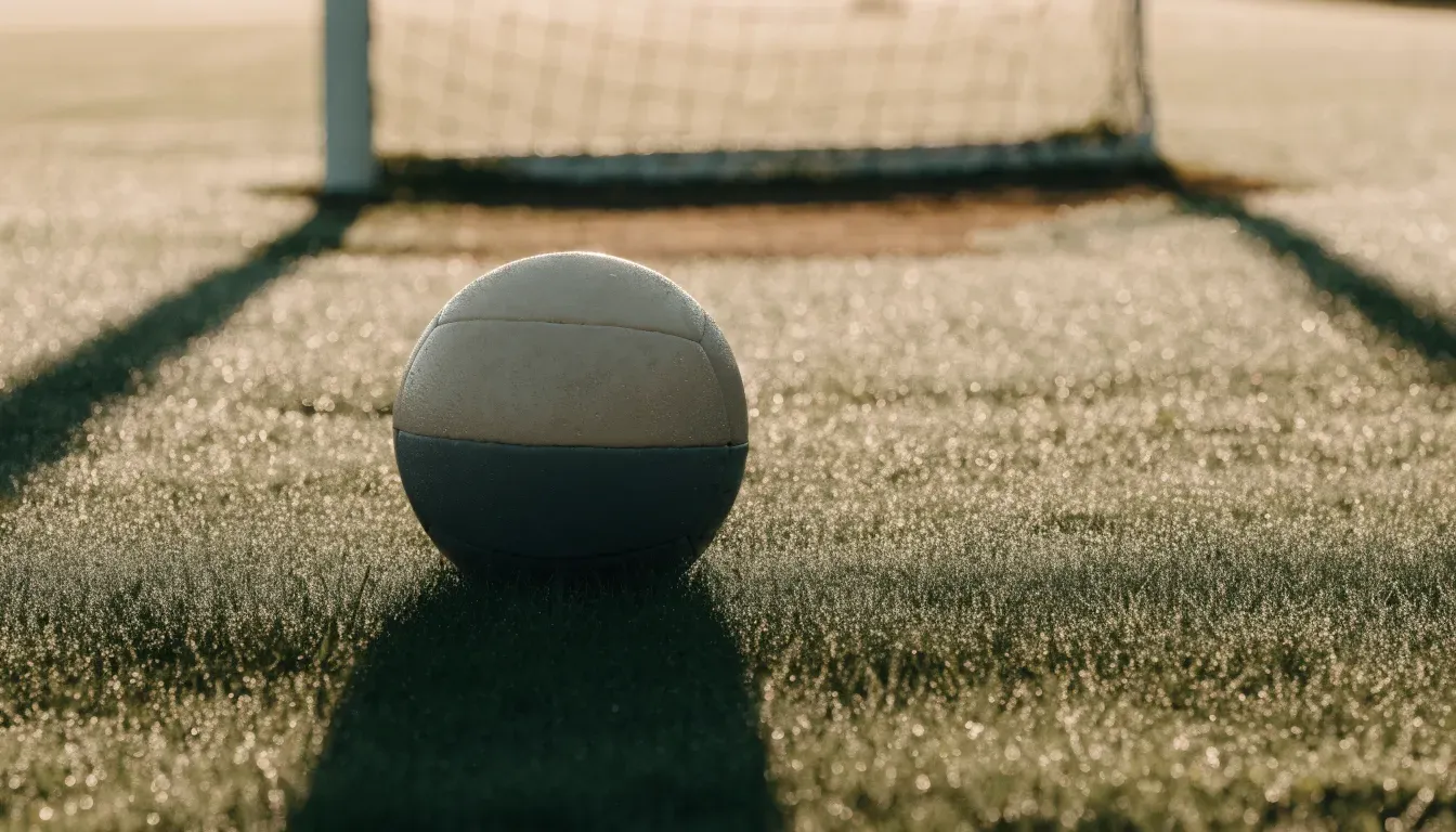 Ein Fussball ruht an der Seitenlinie im taufeuchten Gras, die ersten Sonnenstrahlen fallen schräg über den Platz.