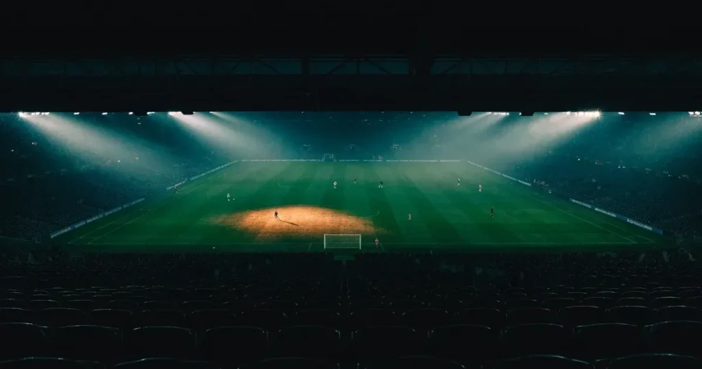 Ein Fussballspiel am Abend aus der Tribünenperspektive, die Spieler sind nur als kleine Figuren zu erkennen und das Flutlicht beleuchtet den nassen Rasen.