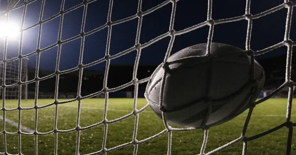 Ein Fussball liegt im Netz eines Tores, Flutlicht und kalte Abendluft liegen über dem leeren Platz
